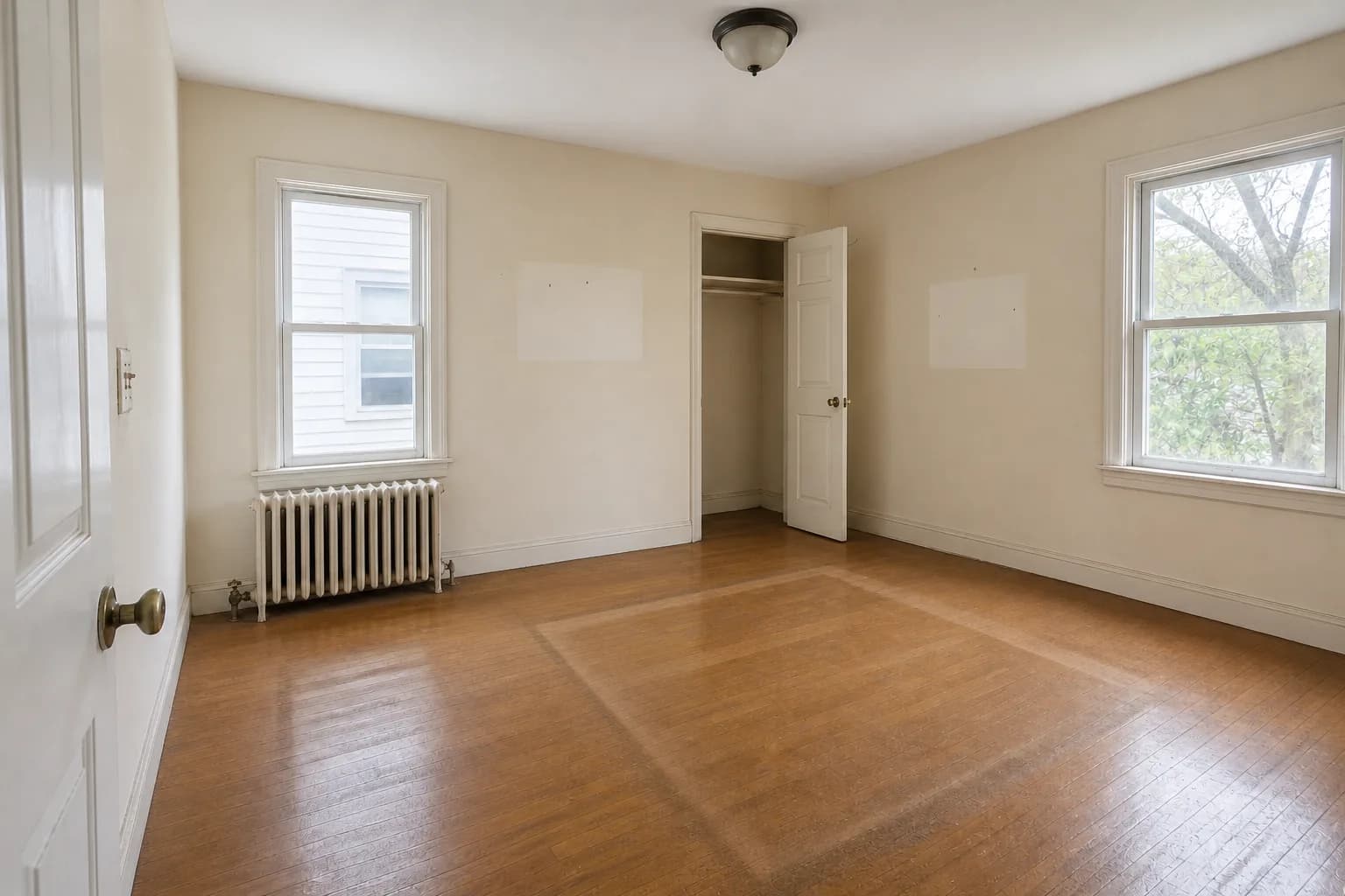 After cleaning: the same emptied room from the same doorway in the same bright late-morning light. The oak floor is swept, vacuumed and mopped so the dust film and lint around the furniture outlines are gone, the dust bunnies are cleared from the baseboards and corners, the scuff and drag marks are scrubbed off the wall, and the hanger and packing paper are removed. The unfaded rectangles where pictures hung and the small nail holes are still there, because cleaning does not repaint.