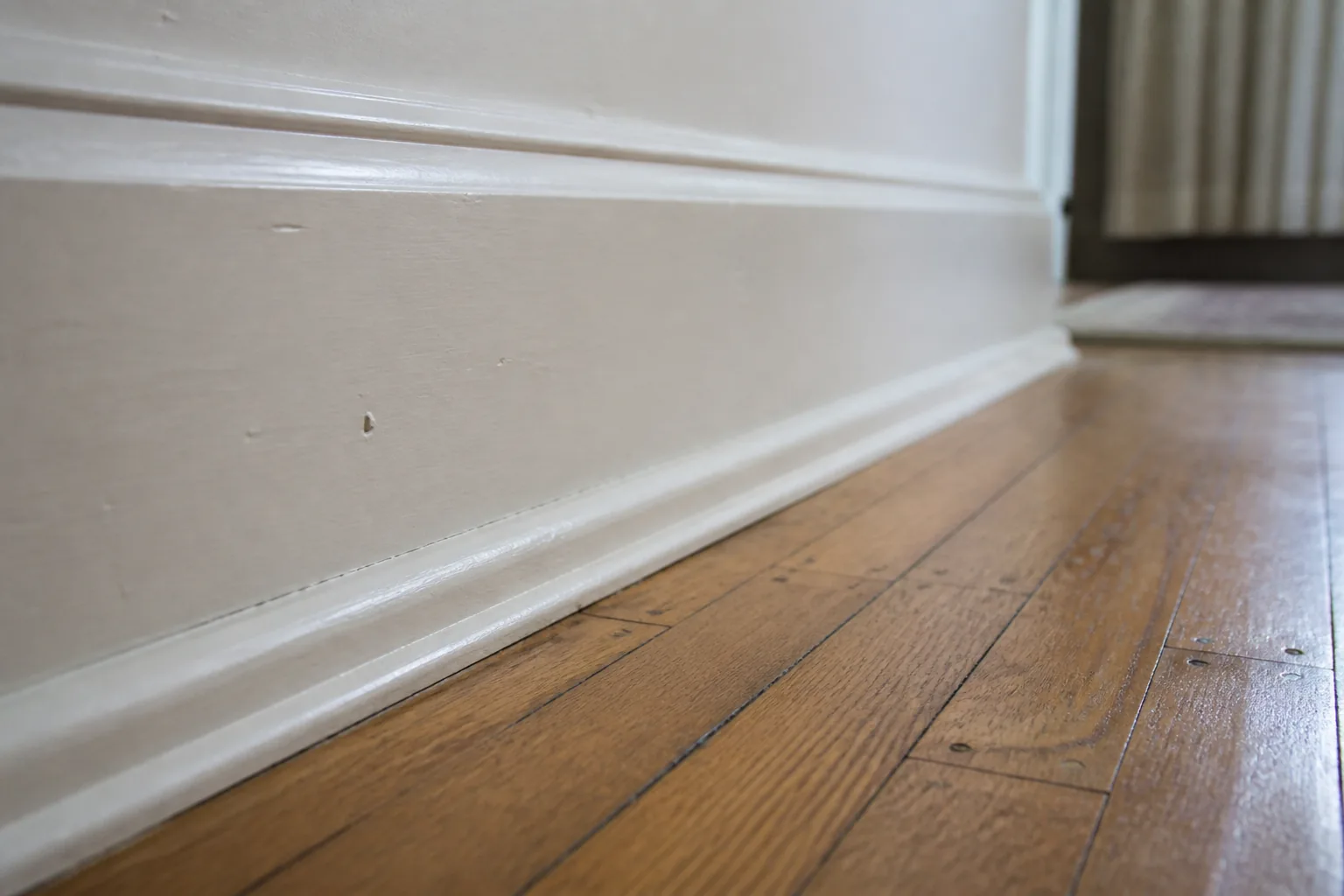 Low raking daylight skimming along a tall painted white baseboard where it meets a worn oak floor, showing the crevice and the floorboards beside it completely free of dust, hair and grey build-up, while the old paint keeps its brush marks and small chips.