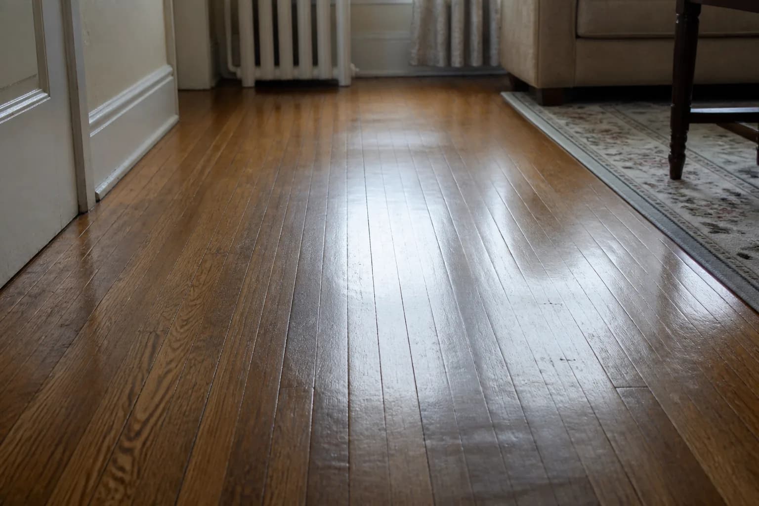 A mopped and dried oak floor seen low and along the boards, the clean satin finish holding a soft stretched reflection of the window above with no cloudy film or streaks, while the wood keeps its darkened grain, old nail holes and worn traffic path.