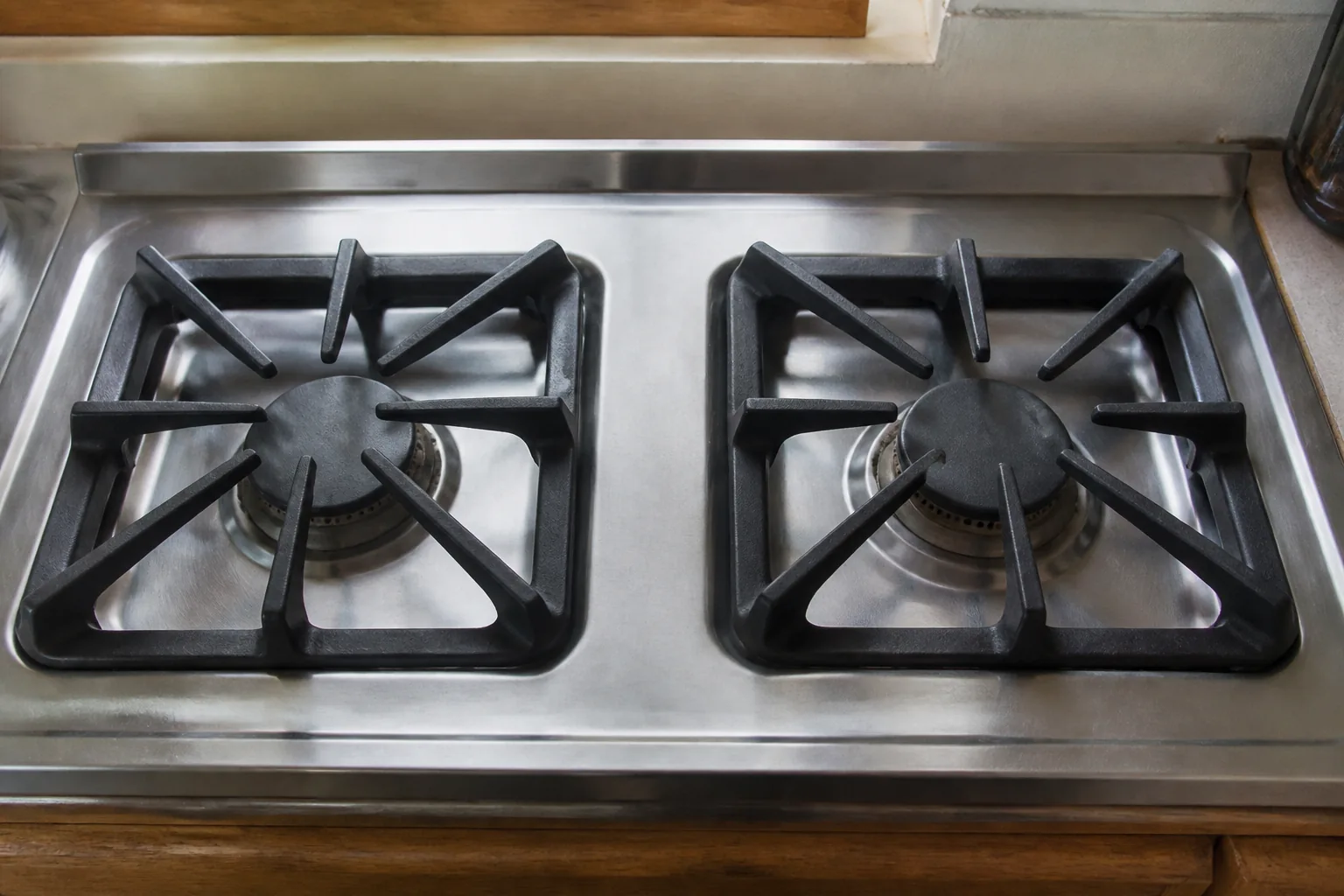 Two heavy cast iron pan supports on a gas cooktop, degreased and scrubbed back to an even matte black with no baked-on spatter, the stainless steel surface between them wiped streak-free, the grates still showing the dull heat discolouration of years of cooking.