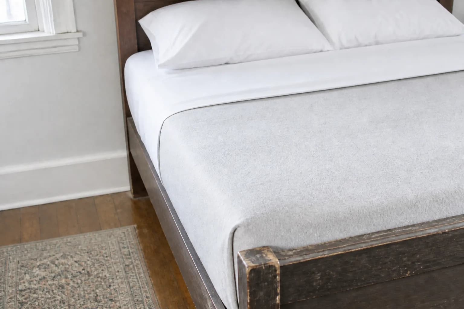 The head of a freshly made bed: a crisp white cotton top sheet turned back in a clean straight fold over a pale grey blanket pulled taut and tucked in, with two plumped pillows against a dark wooden headboard and the old bed frame showing its wear.