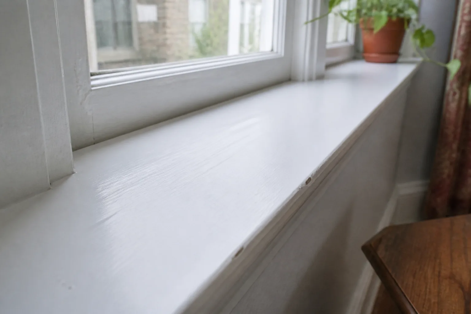 Daylight raking along a painted white wooden windowsill in an older house, dusted right into the corner where the sill meets the sash with no grey build-up left, a small terracotta pot of a trailing plant at the far end, and the old paint showing its brush marks and chips.