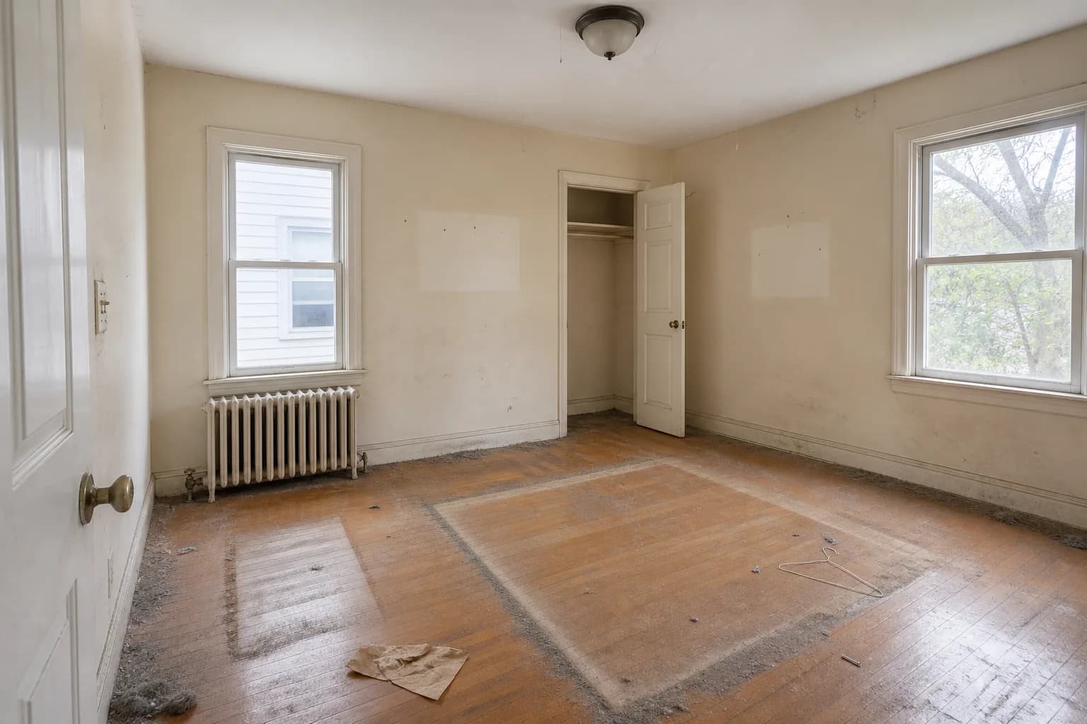 Before cleaning: an emptied room with cream walls and an oak floor, cleared for a move. Grey dust and lint outline the rectangles where a bed and dresser stood, dust bunnies bank along the baseboards and corners, dark scuff and drag marks streak the lower wall, unfaded rectangles and nail holes mark where pictures hung, and a wire hanger and a sheet of crumpled packing paper are left on the floor.