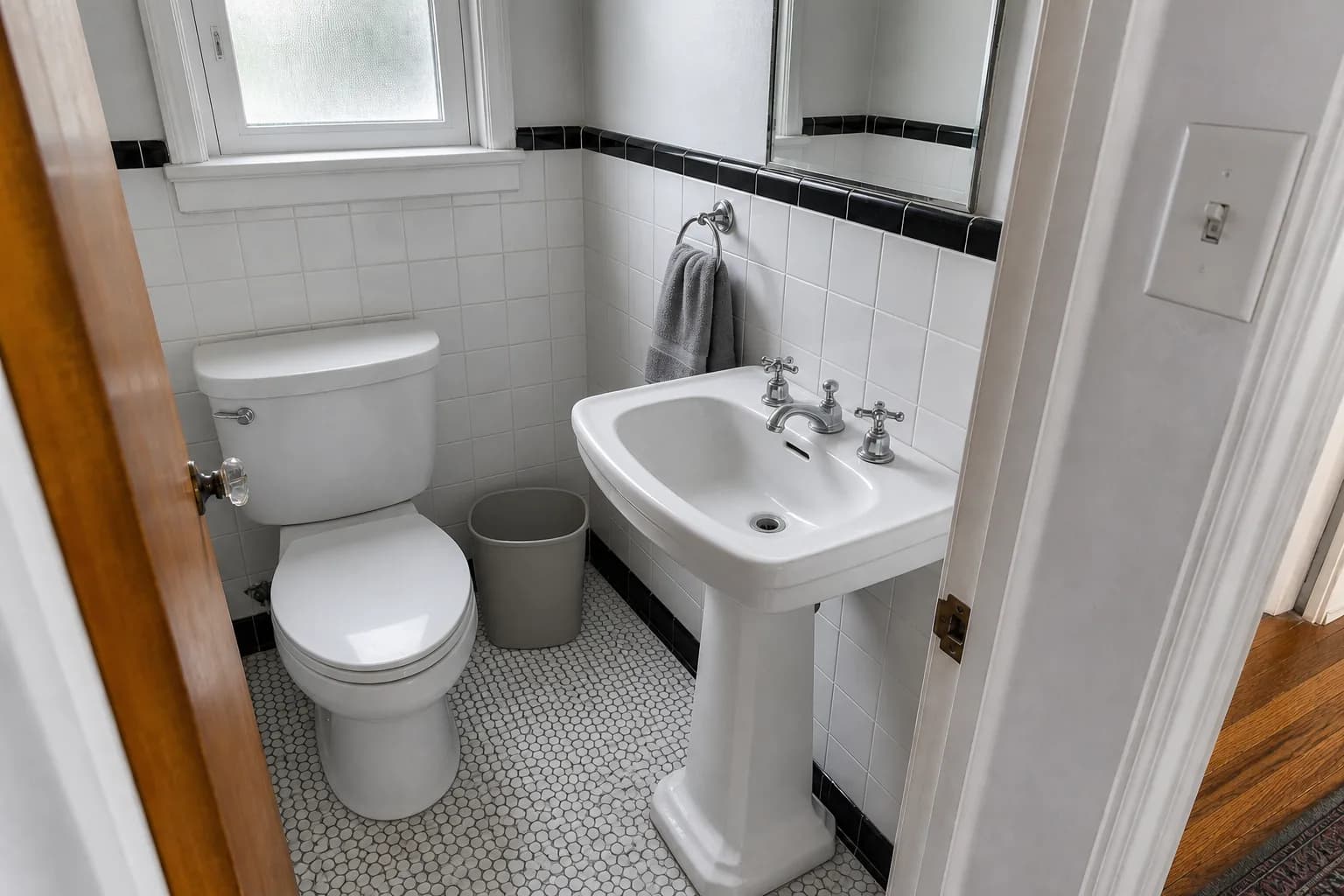 After cleaning: the same powder room from the same doorway in the same flat light. The hard-water scale is dissolved off the spout and both cross handles so the chrome is polished bright, the mirror is spotless with the toothpaste spatter gone, the sink bowl is white with the grimy ring and the rust-brown mineral streak removed, the toilet is cleaned inside and out and the tank top dusted, the switch plate is wiped, the hand towel is folded on its chrome ring, the bin is empty and the penny tile floor is mopped so the grout reads noticeably lighter. The tile keeps its fine vintage crazing.